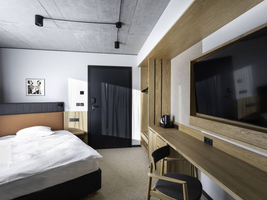 A modern hotel room with a bed, a desk, a chair, a large TV, and a door leading to another room.