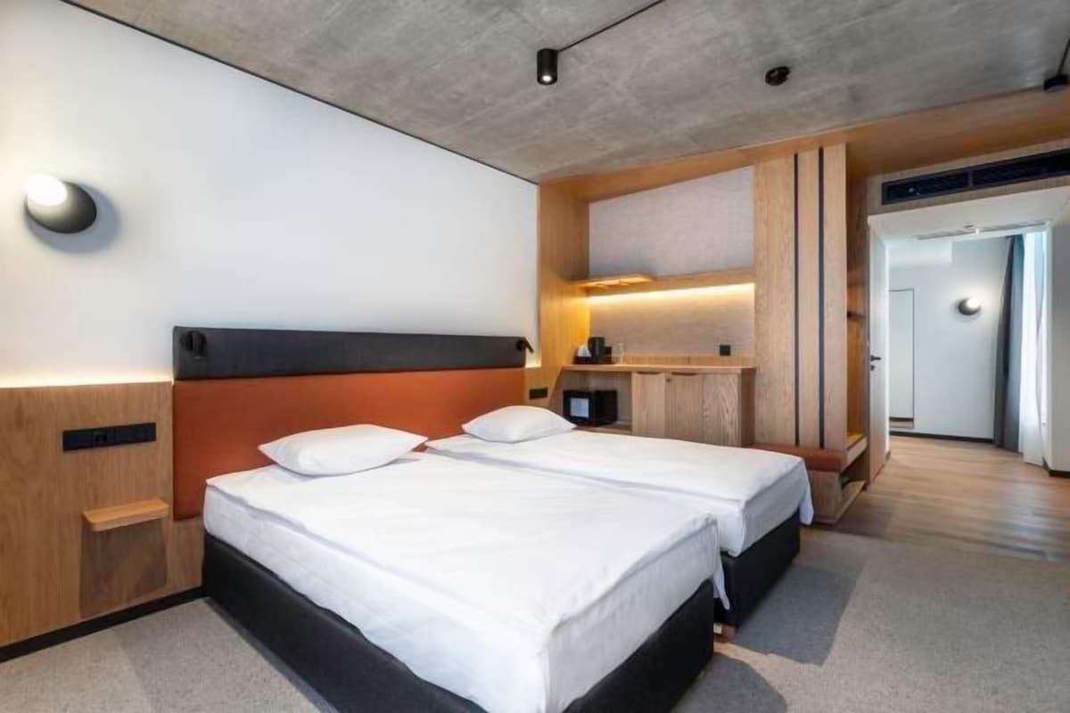 A modern hotel room with two beds, a wooden headboard, a built-in desk, and a small seating area.