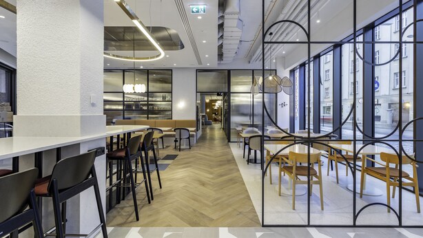 Un café moderno con una barra larga, taburetes y mesas. Cuenta con amplios ventanales en un lateral y vistas a la calle.
