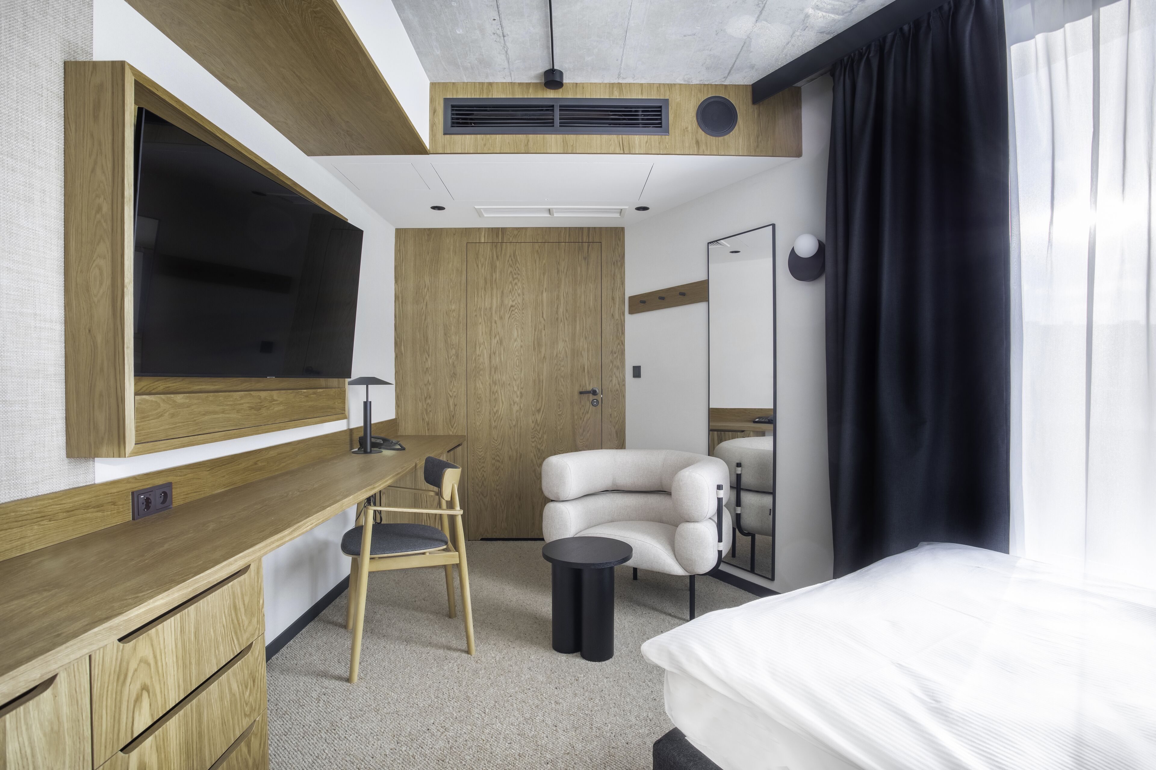 A modern hotel room with a wooden desk, a chair, a bed, and a large mirror.