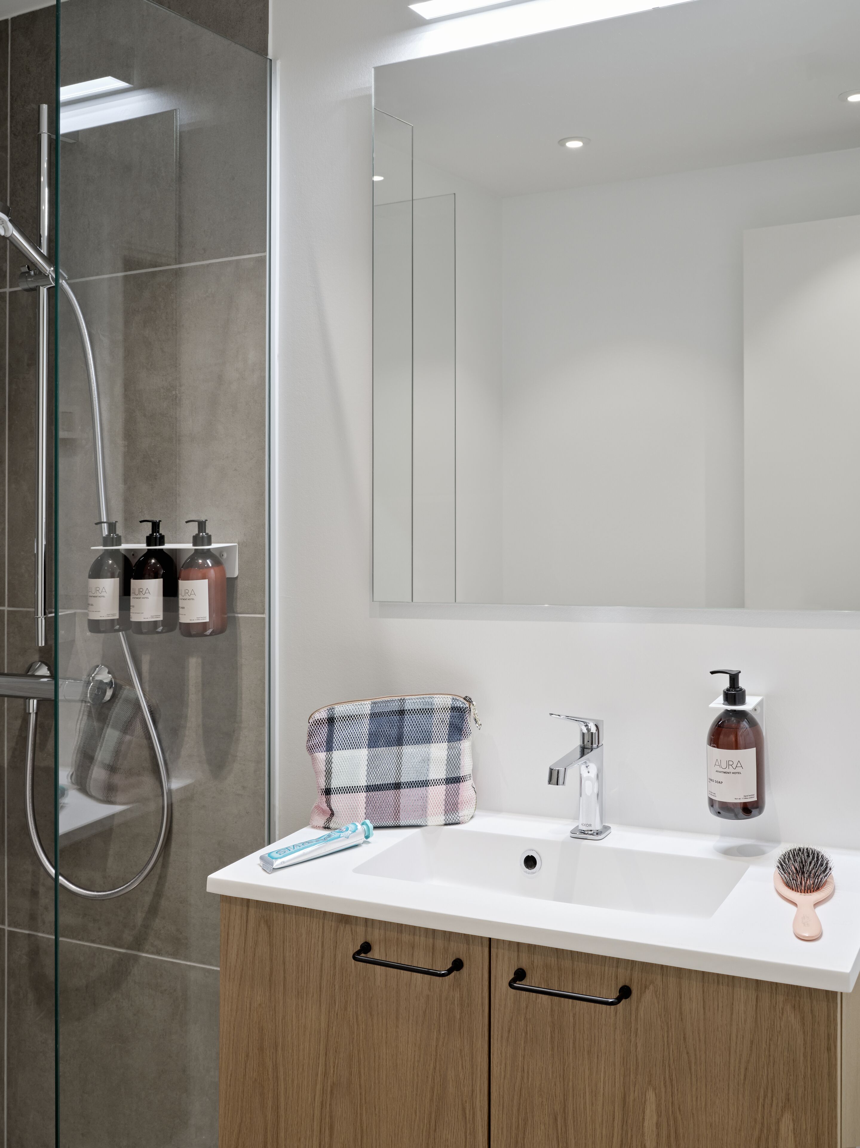 A modern bathroom with a glass-enclosed shower, a white sink with a mirror above, and toiletries on a shelf.