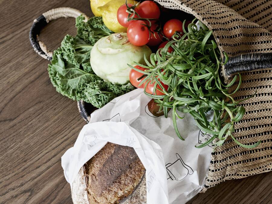 A woven bag containing fresh vegetables and a loaf of bread on a wooden table.