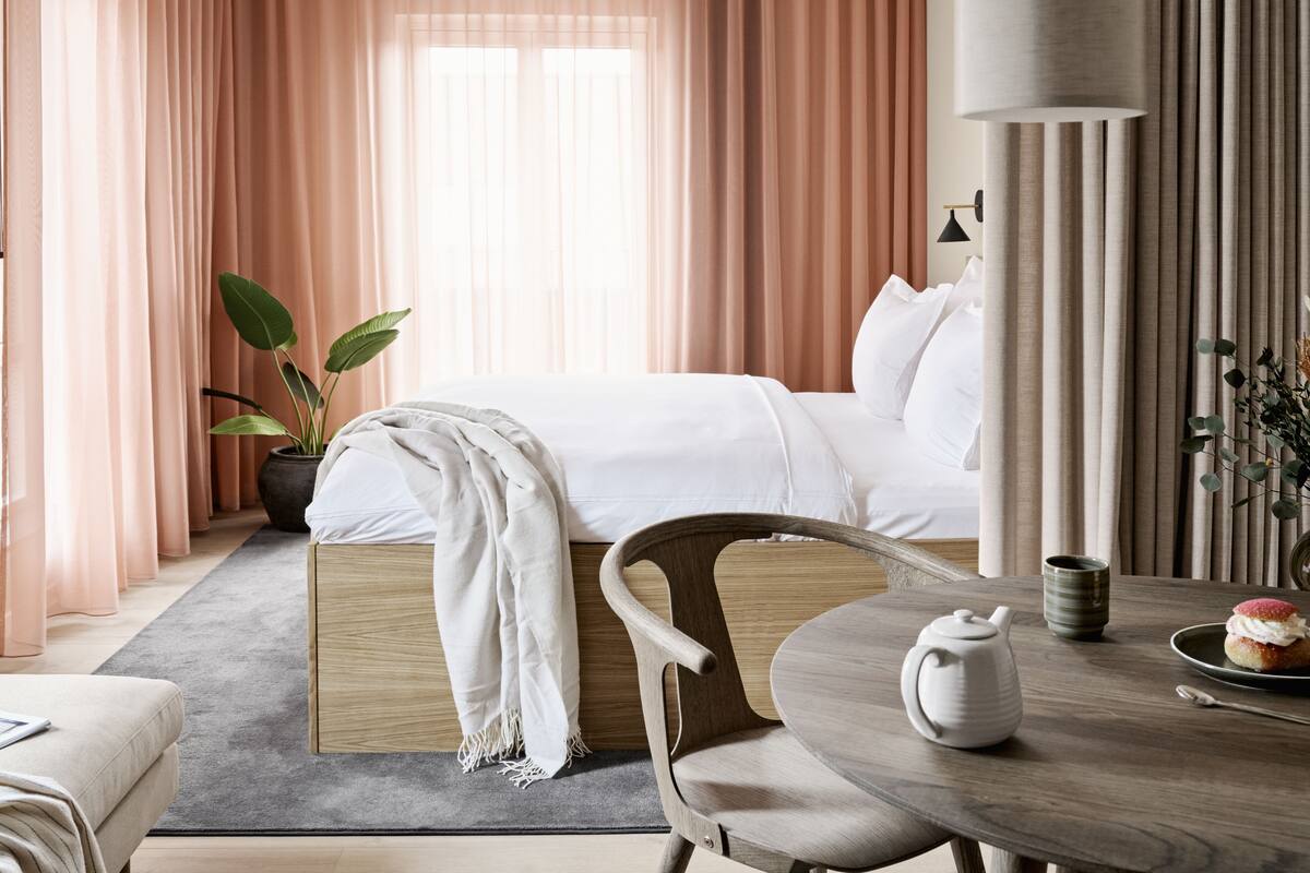 A hotel room with a bed, a chair, a table with a teapot and a plate of food, a potted plant, and curtains.