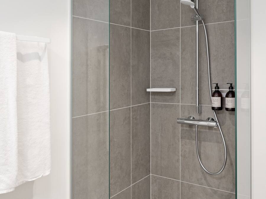 A walk-in shower with a glass door, grey tiles, a handheld showerhead, a wall-mounted faucet, and two bottles of shampoo on a shelf.