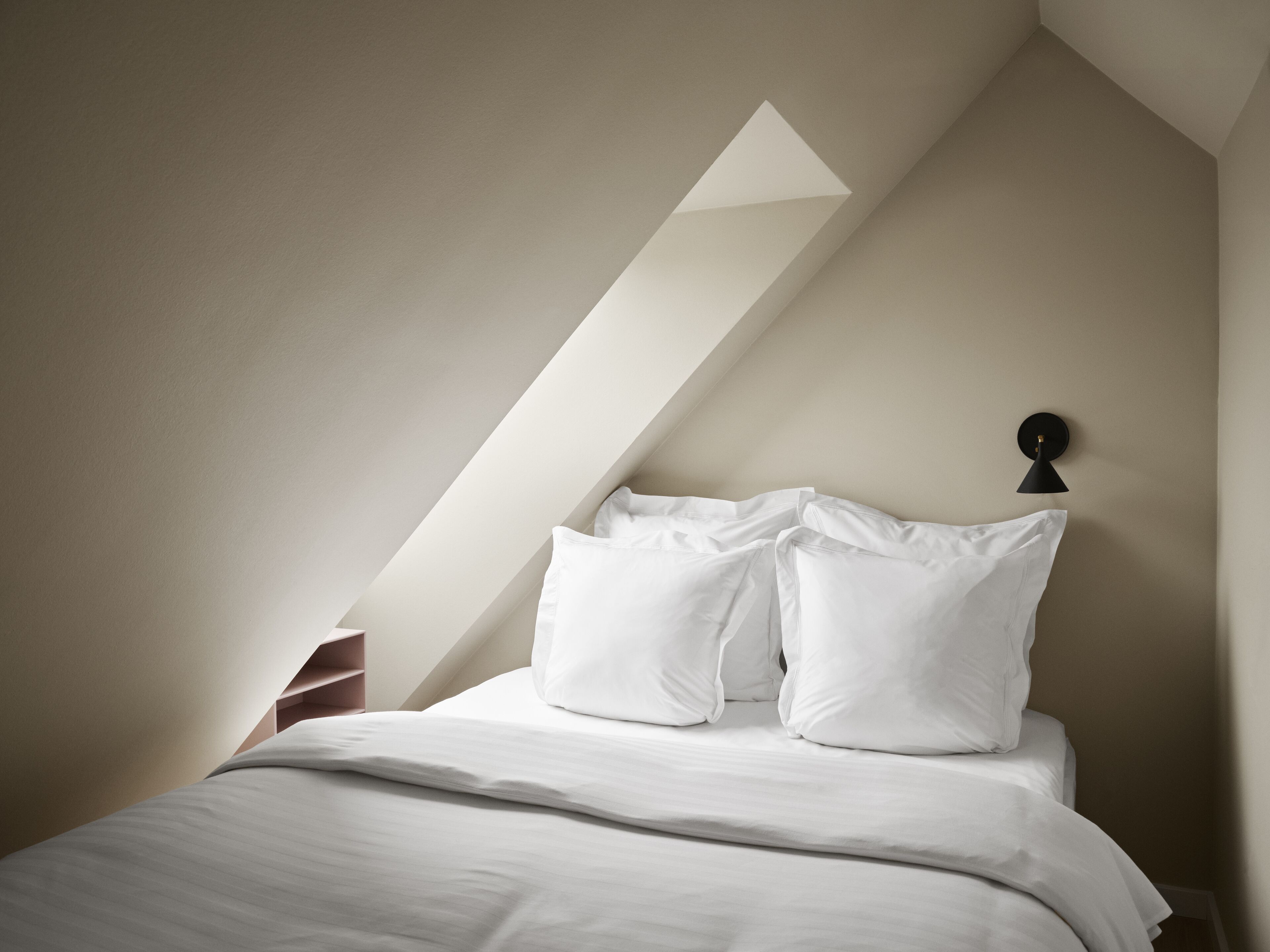 A bedroom with a sloped ceiling, a bed with white bedding, and a wall-mounted lamp.