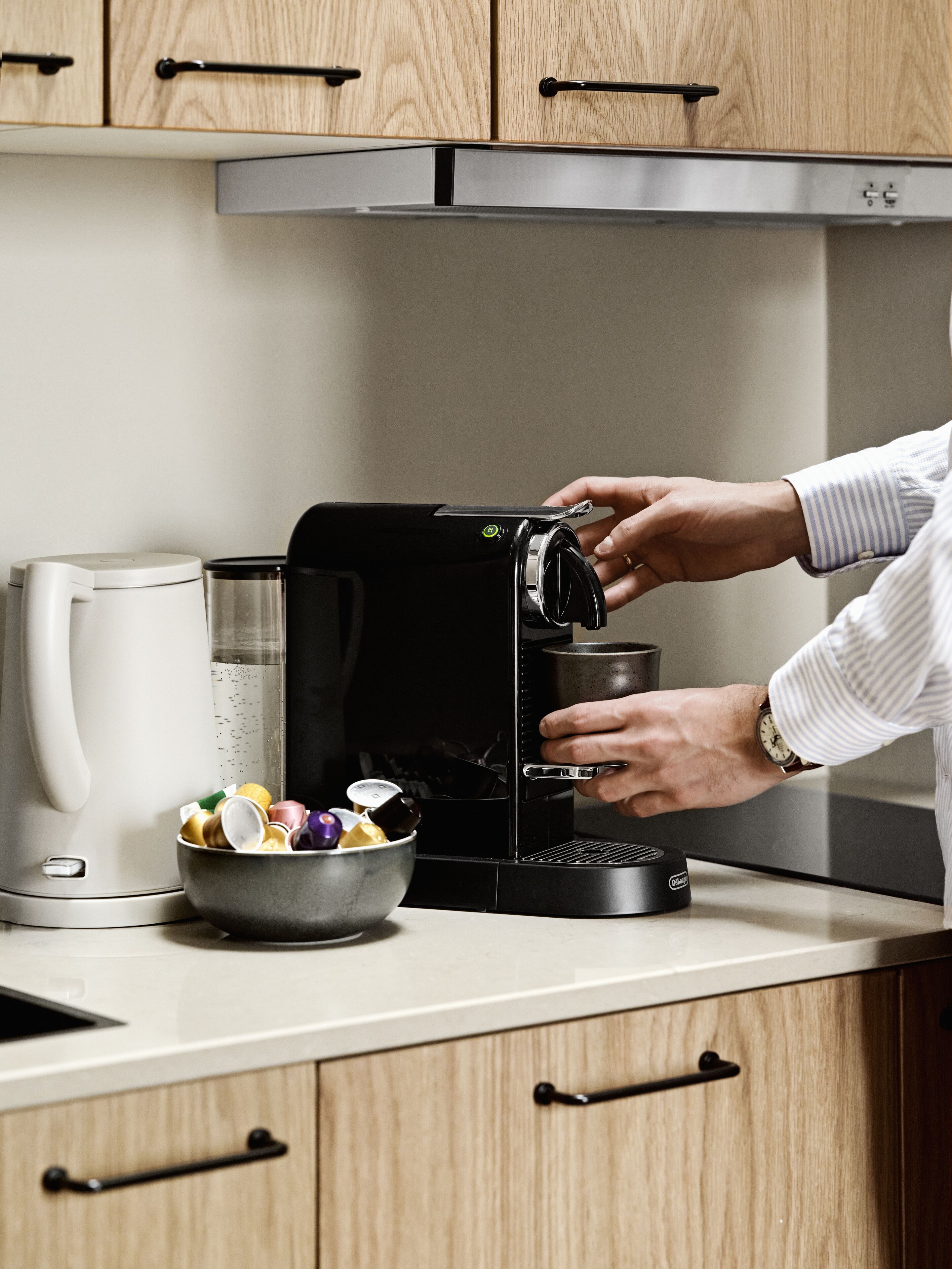 Una cocina con una cafetera, un bol de cápsulas de café y una persona preparando café.