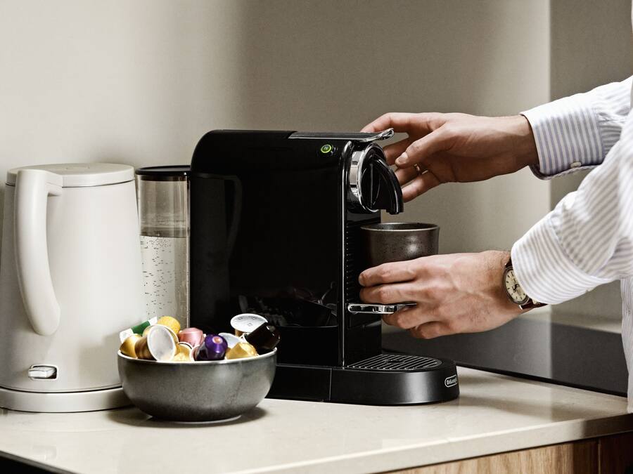 Una cocina con una cafetera, un bol de cápsulas de café y una persona preparando café.