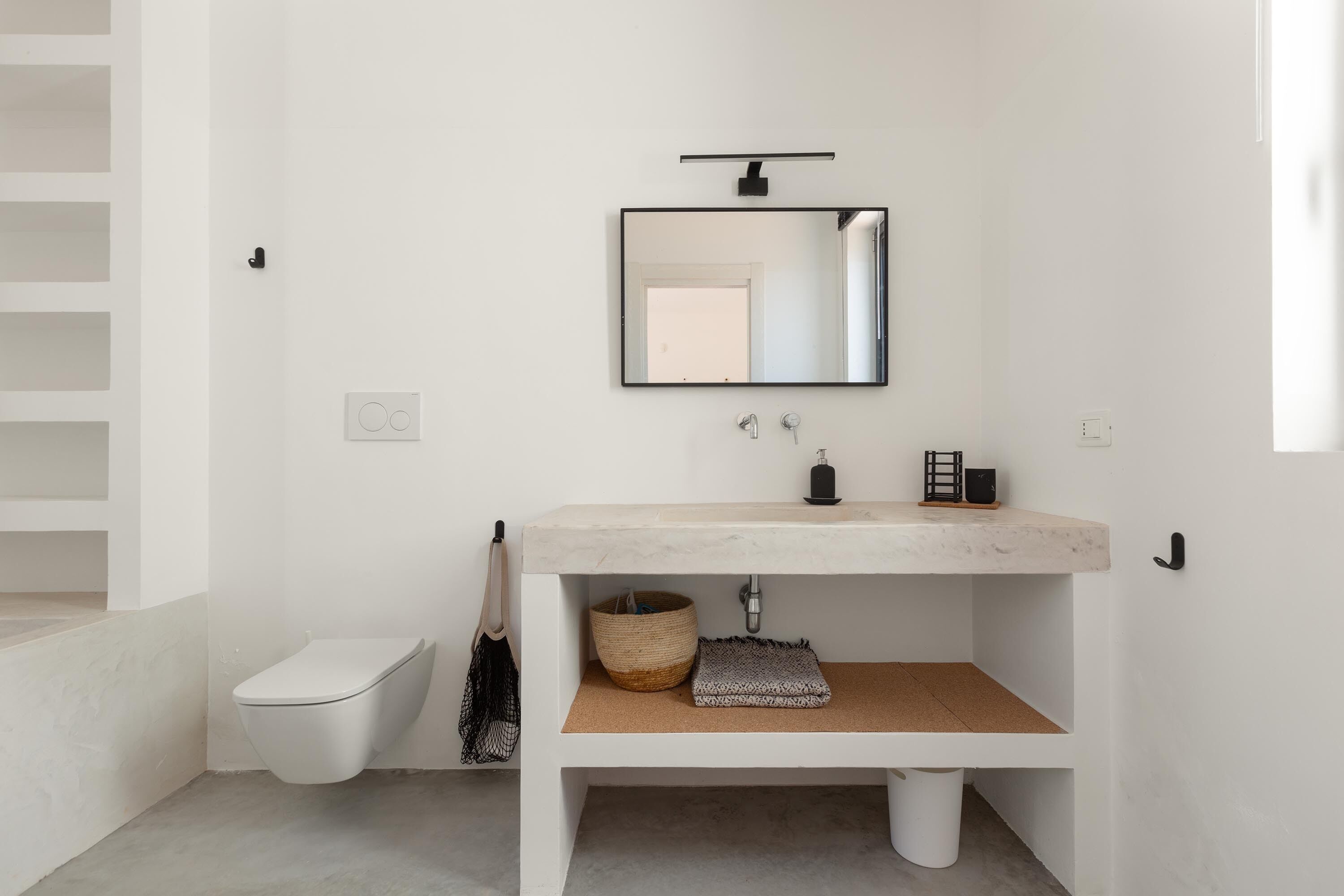 Un baño moderno con un lavamanos blanco, un espejo, un inodoro y un pequeño estante con una cesta y una toalla.