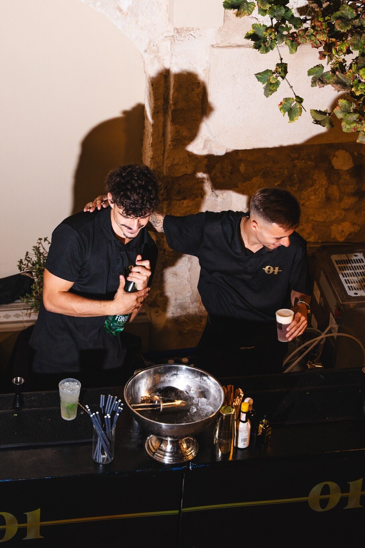 Dos personas preparando bebidas en un bar, con un bol de metal, hielo y cocteleras.