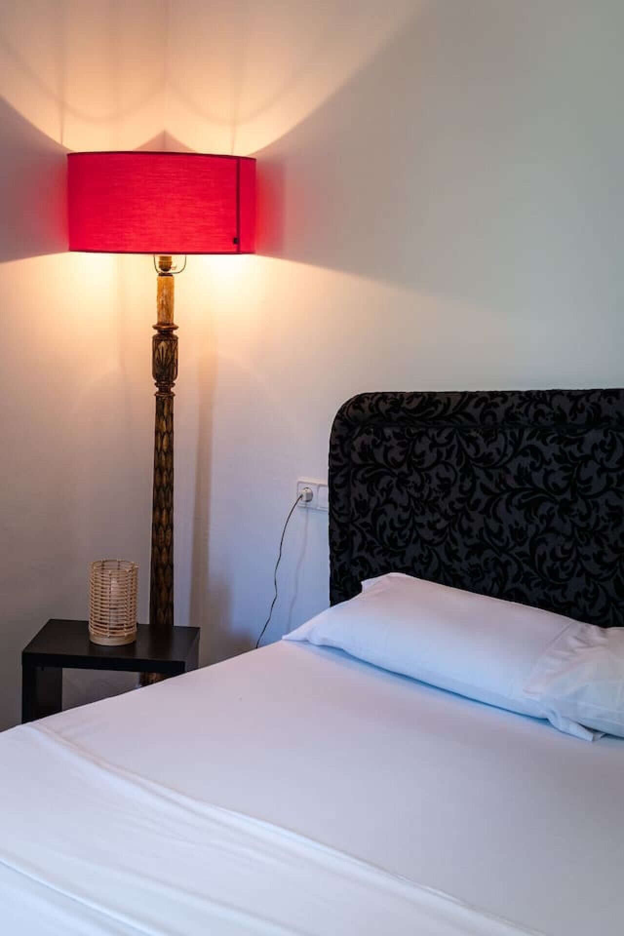 Un lit bien fait, avec du linge de lit blanc, une lampe rouge sur une table de chevet et une tête de lit à motifs.