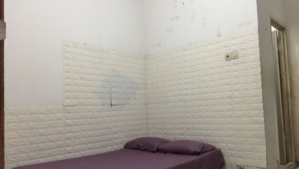 A small, narrow room with white brick walls, a single bed with a purple mattress, and a door on the right side. - OYO Life 93009 Griya Nautika Sedati (Sidoarjo)