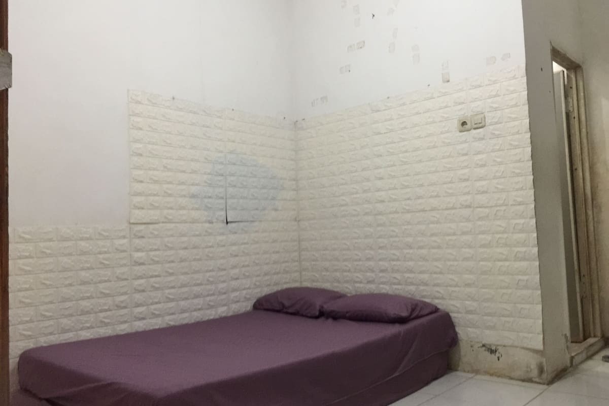 A small, narrow room with white brick walls, a single bed with a purple mattress, and a door on the right side.