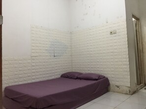 A small, narrow room with white brick walls, a single bed with a purple mattress, and a door on the right side. - OYO Life 93009 Griya Nautika Sedati (Sidoarjo)