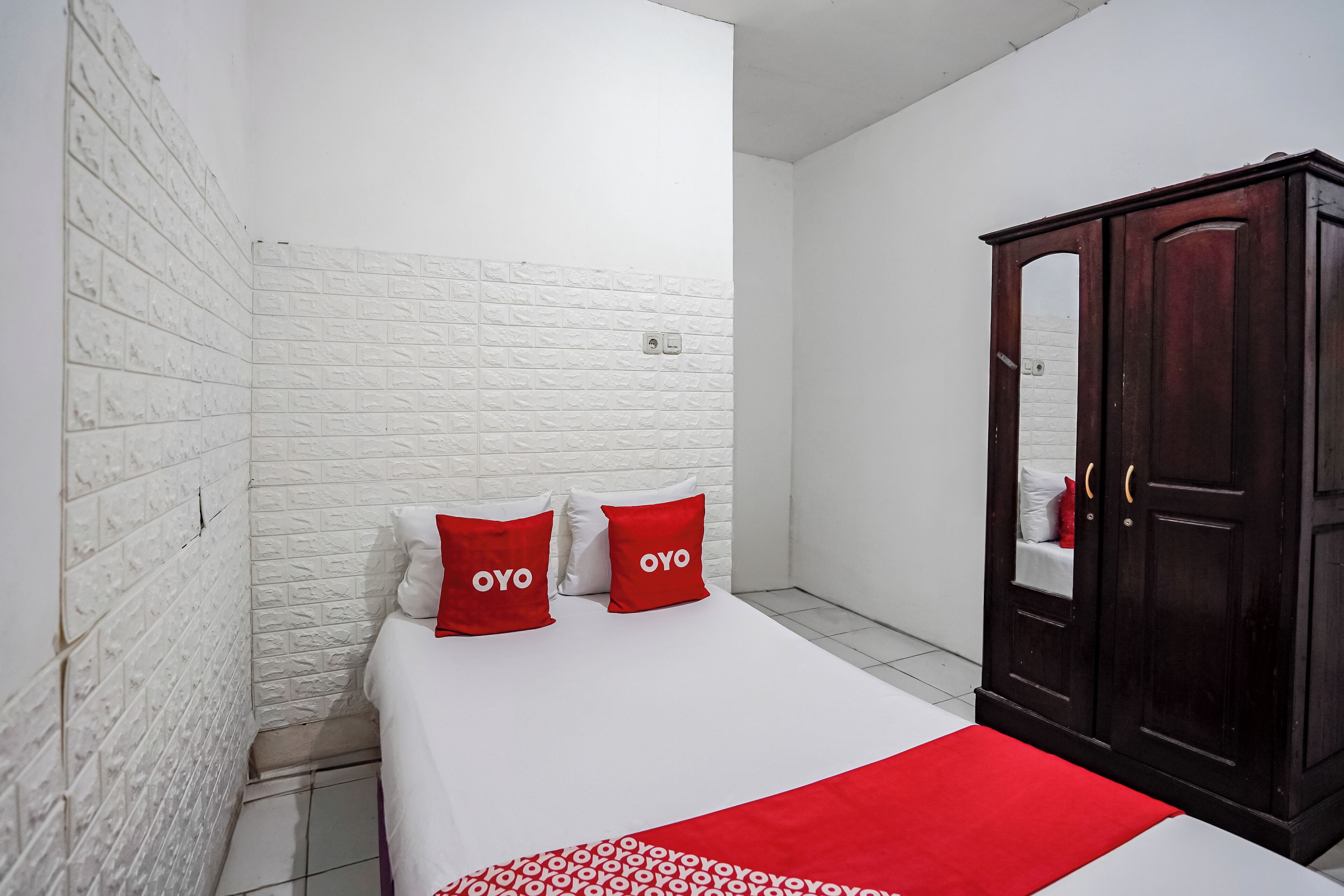 A bedroom with a white brick wall, a wooden wardrobe, and two red pillows with the word 'OYO' on them.