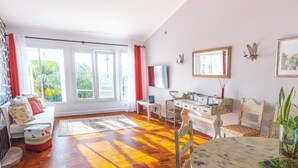Ein geräumiges Wohnzimmer mit Fischgrat-Holzboden, einem Esstisch mit Stühlen, einer Couch mit Kissen, einem Fernseher und einem großen Fenster mit Vorhängen.