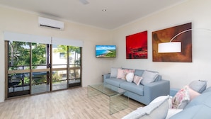 A modern living room with a glass coffee table, a grey sofa with decorative pillows, and a large sliding glass door leading to a balcony with a view of greenery and a building.