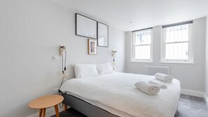Un dormitorio moderno con una cama grande, una mesita de madera y dos ventanas que dejan entrar luz natural.
