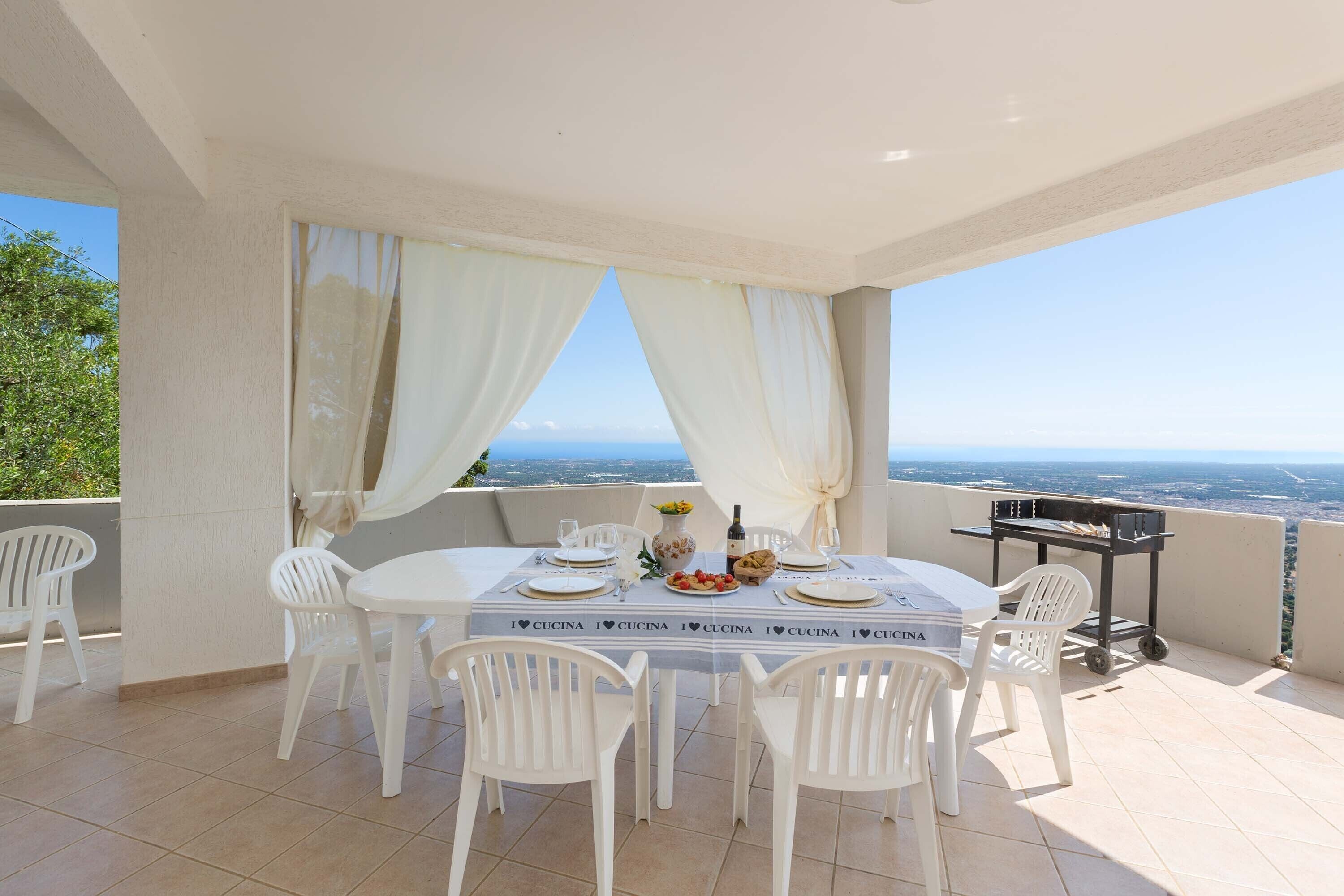 A dining area with a table set for four, white chairs, a grill cart, and a view of the landscape.