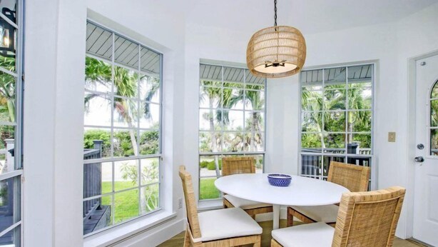 A dining area with a round table and wicker chairs, large windows with a view of greenery, and a hanging light fixture. - One More Day (Sanibel)