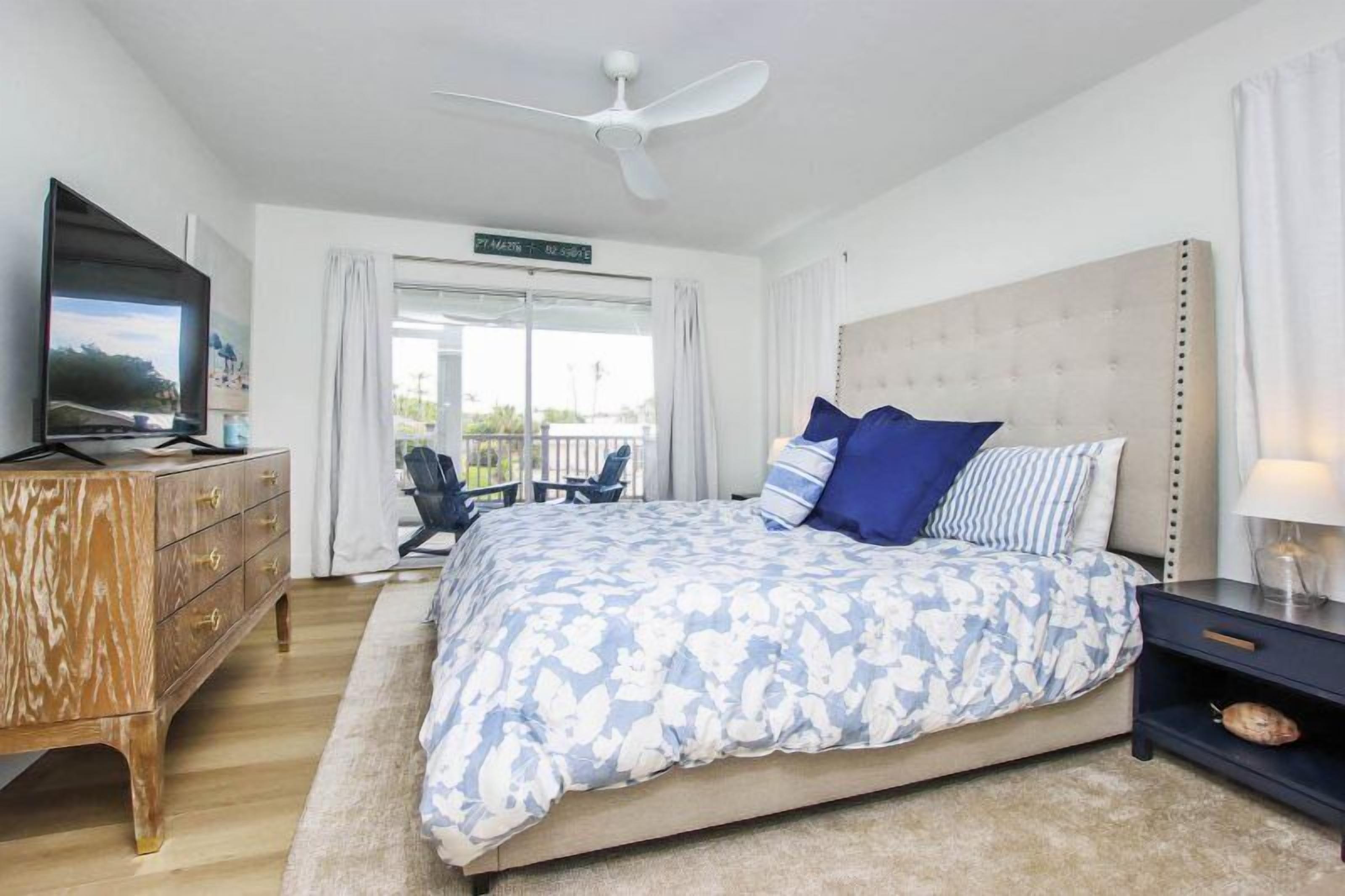 A bedroom with a large bed, a TV, a ceiling fan, a dresser, and a balcony with a view.