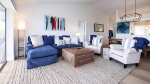 A modern living room with a sectional sofa, a coffee table, and a white armchair. There is a painting on the wall and a hanging light fixture. - One More Day (Sanibel)