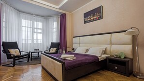 A hotel room with a large bed, two chairs, a nightstand, a lamp, and a cityscape painting on the wall. - Stylish apartment near the railway station (Minsk)