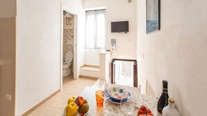Una habitación pequeña y luminosa con una mesa de comedor dispuesta con frutas, una botella y un vaso. Hay un televisor montado en la pared y se ve una ventana con cortinas.