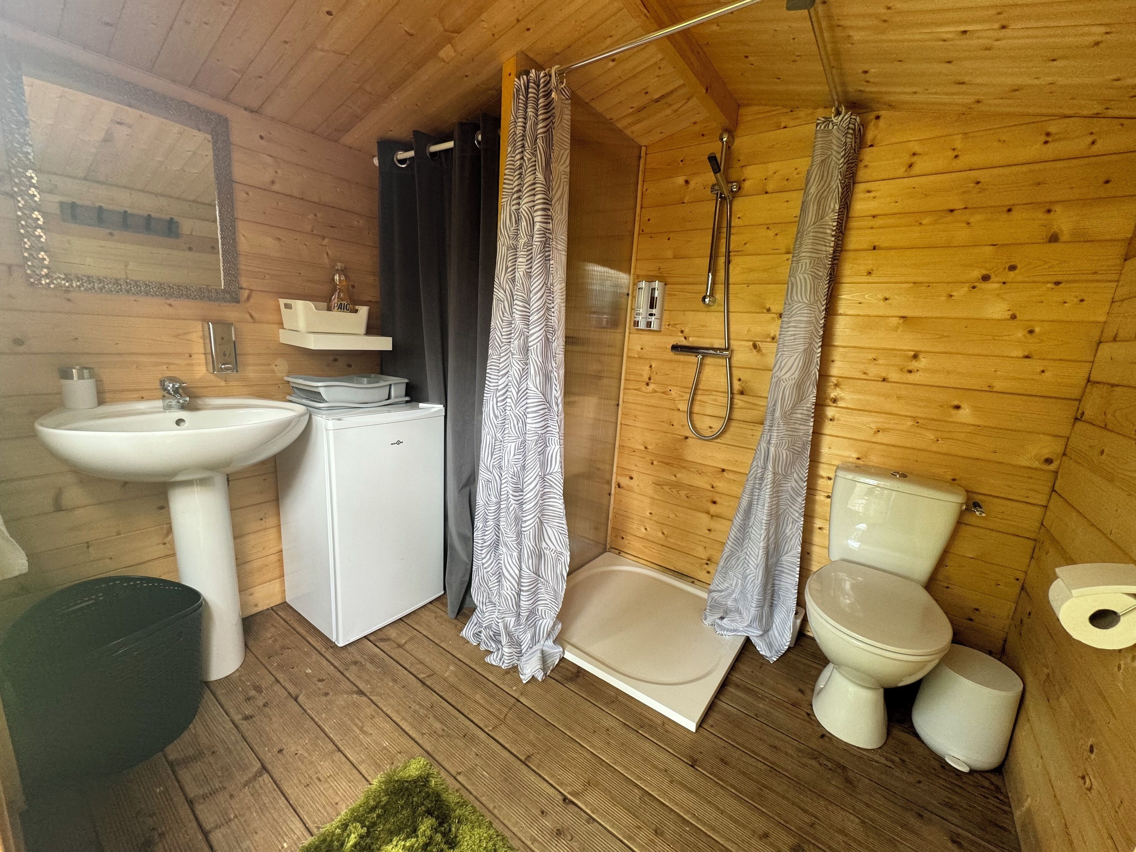 Et lille, træbeklædt badeværelse med vask, toilet, bruser og et minikøleskab.