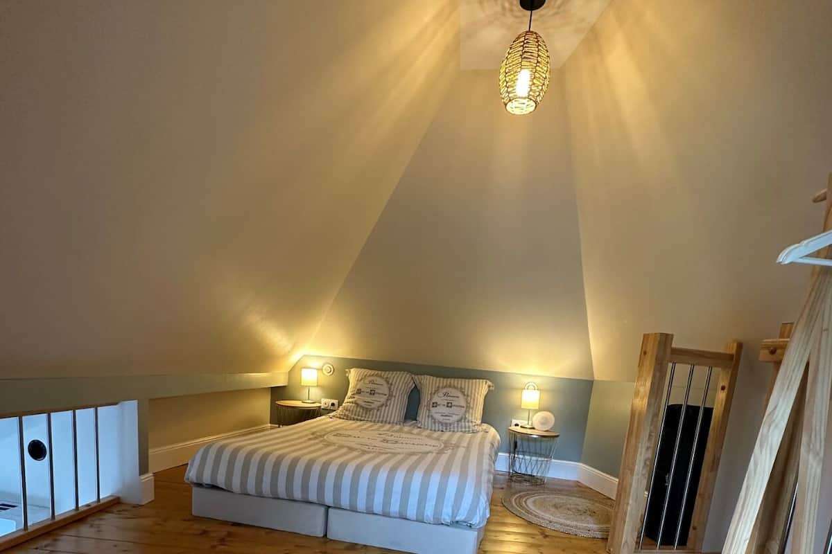 Une chambre mansardée chaleureuse, avec un plafond en pente, un lit avec une literie à rayures, deux lampes de chevet et un luminaire suspendu.
