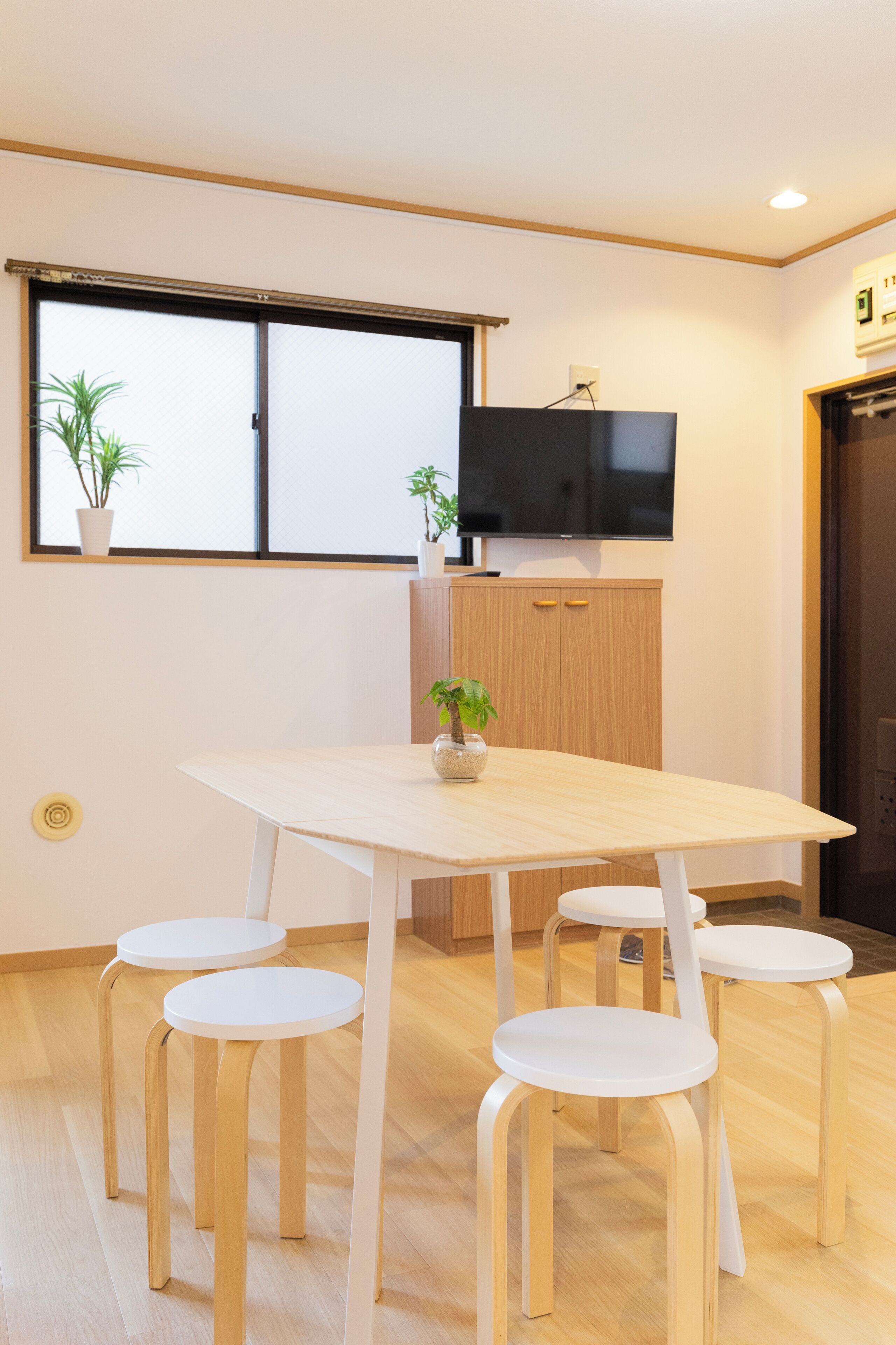 A modern kitchen with a wooden table and four white chairs, a TV mounted above the cabinets, and two potted plants on the windowsill.