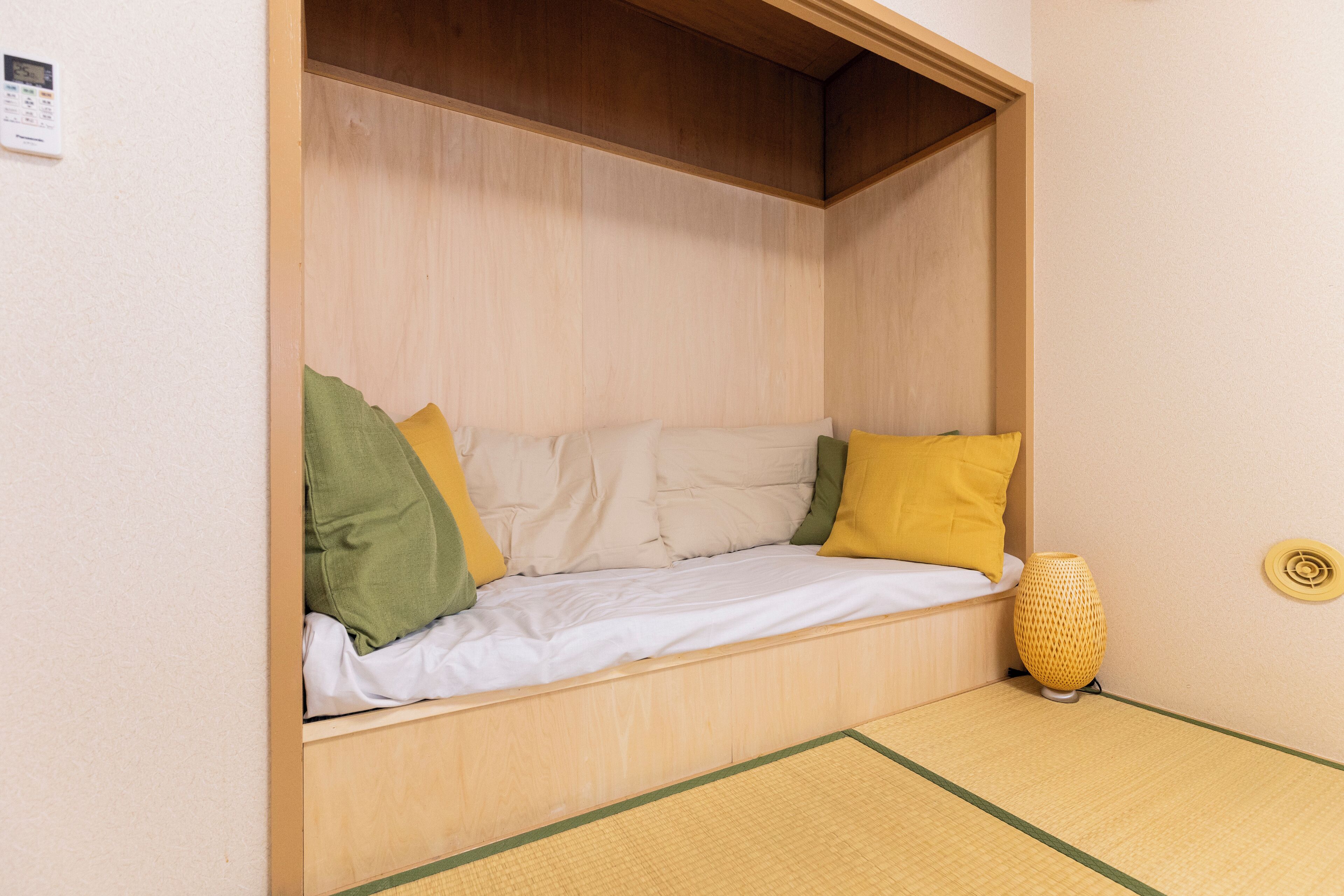 Una stanza accogliente e piccola, con un letto di legno, un cuscino verde e giallo e una lampada gialla.