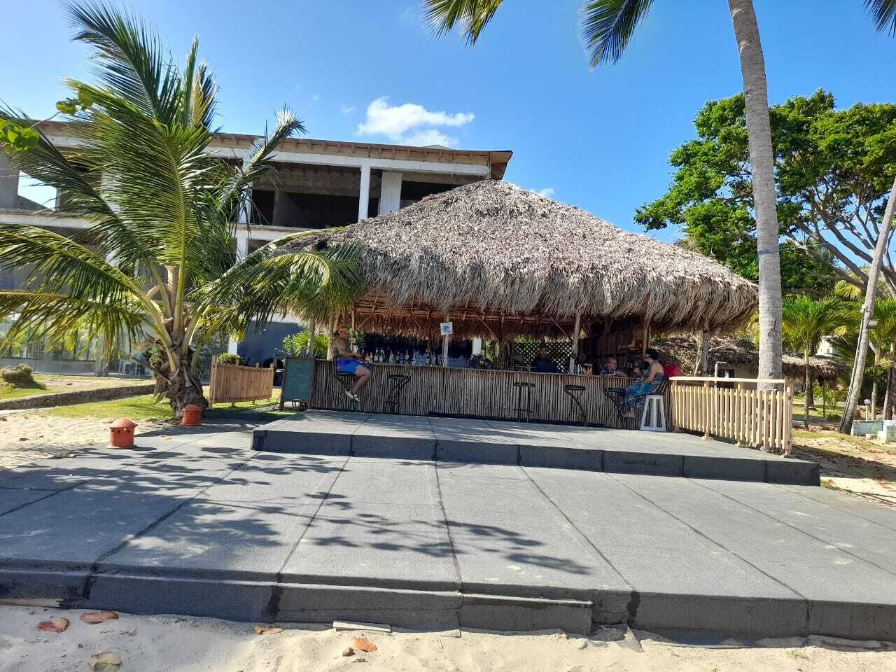 A beachside bar with a thatched roof, palm trees, and a concrete walkway.