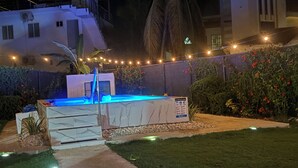 A well-lit outdoor pool area with a clear pathway leading to it, surrounded by a lush garden and a modern building in the background.