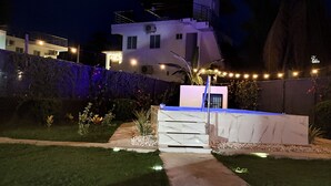 A modern house with a white exterior, a balcony, and a well-lit garden area at night.