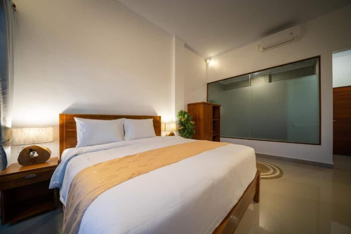 A modern hotel room with a large bed, a nightstand with a lamp, a wooden wardrobe, and a large mirror.