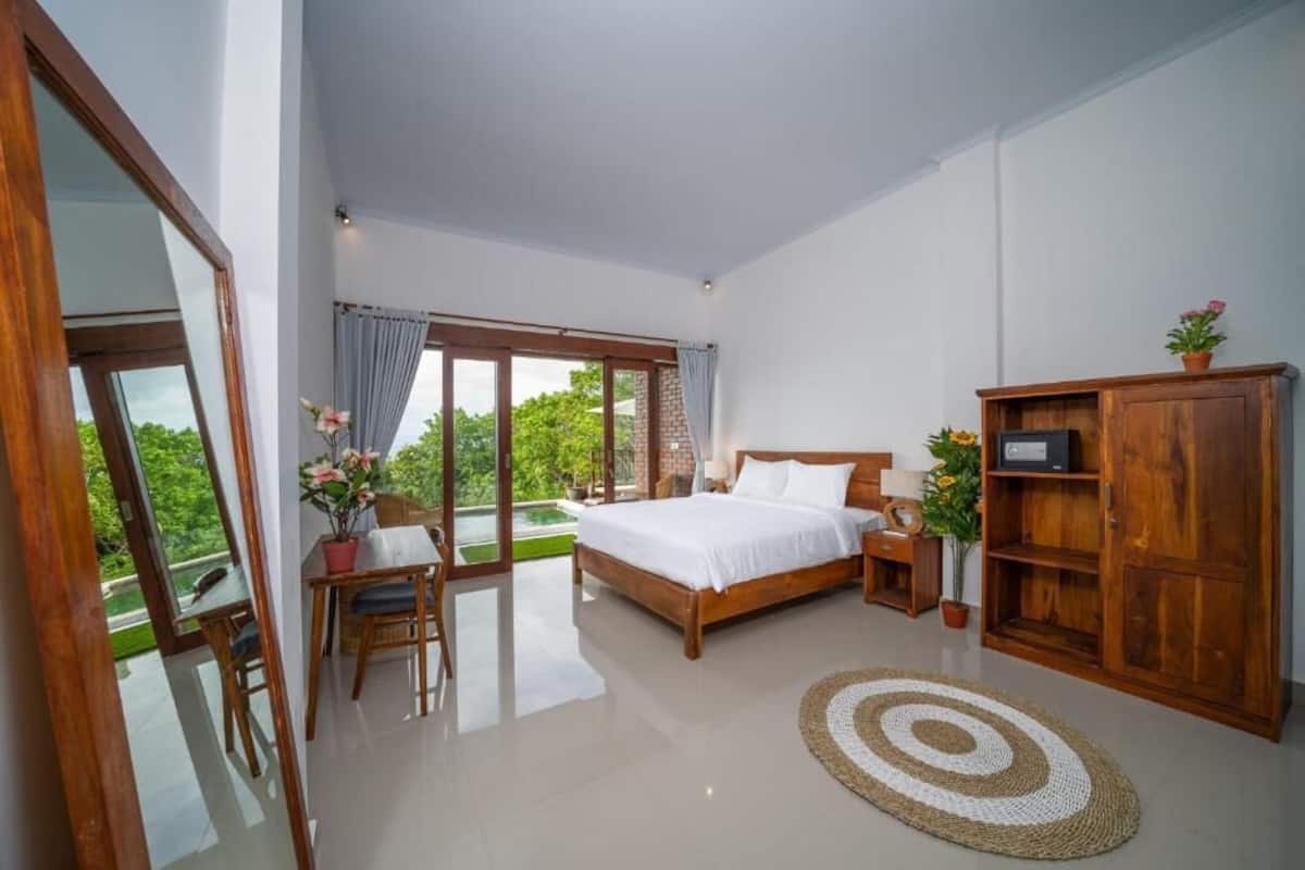 A bedroom with a large bed, a wooden wardrobe, a desk with a chair, and a view of greenery through the window.