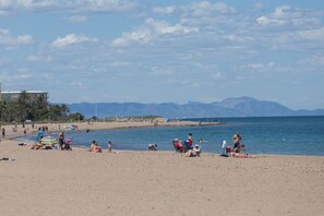 Una playa con gente tomando el sol y nadando, una sombrilla colorida y montañas al fondo.