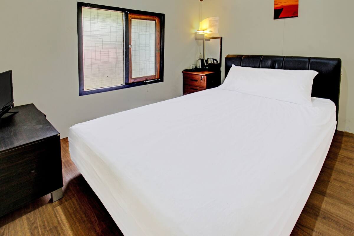 A neatly made bed with white linens, a wooden nightstand with a lamp, and a window with blinds.
