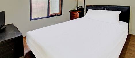 Uma cama bem arrumada com lençóis brancos, um criado-mudo de madeira com uma luminária e uma janela com persianas.
