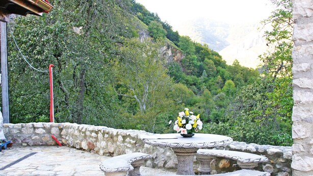 A stone patio with a table and benches, a vase of flowers, and a mountainous backdrop. - Apartamentos Valdecarzana . Braña el Revellón Senda del oso (TEVERGA-OVIEDO-ASTURIAS)