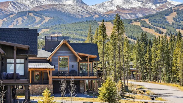 Une maison moderne à deux étages, dotée d’un balcon, entourée d’une forêt et avec des montagnes en arrière-plan.