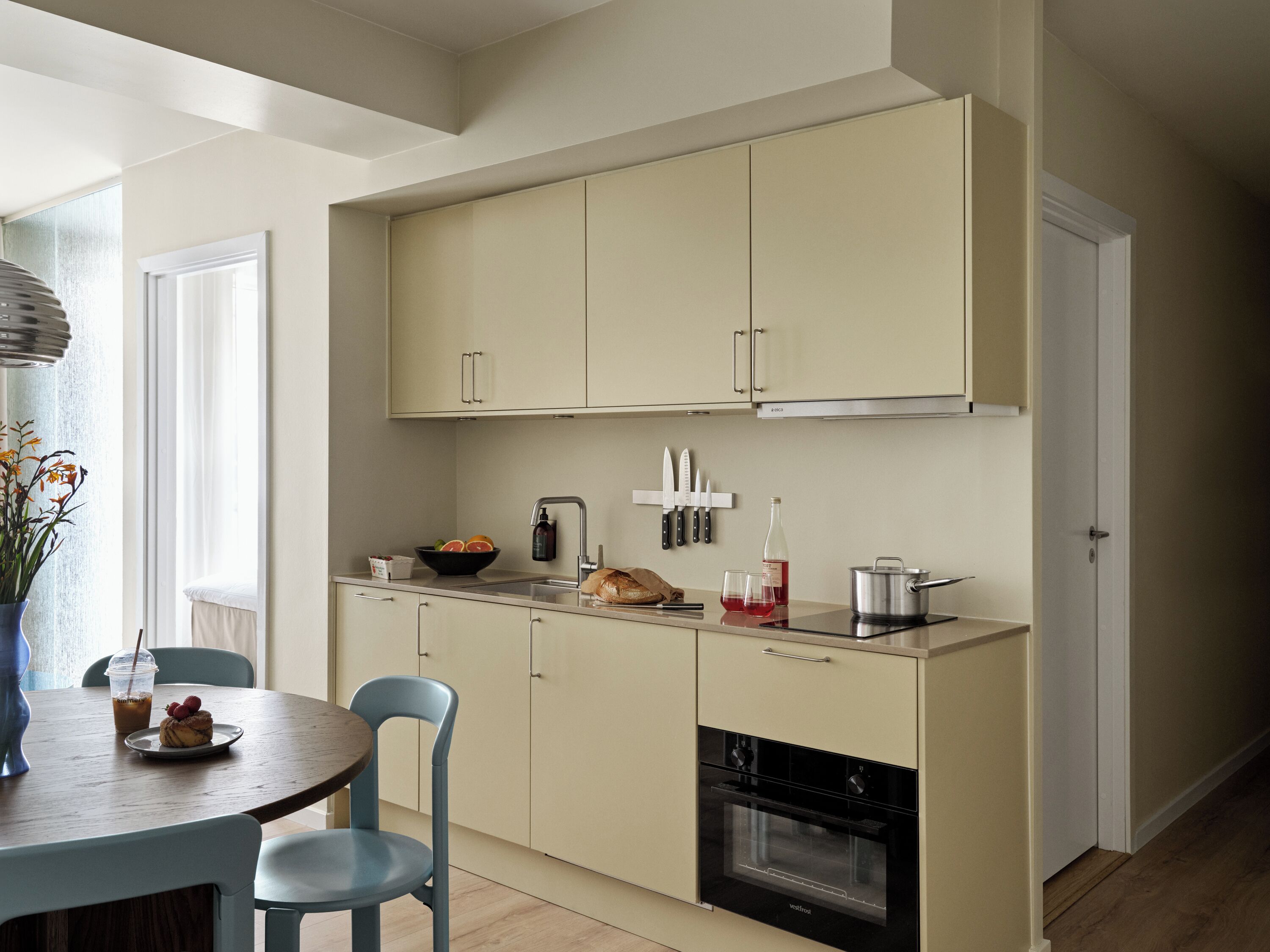 Een moderne keuken met lichte kastjes, een eettafel met stoelen, een inbouwoven en een raam met uitzicht op de omgeving.