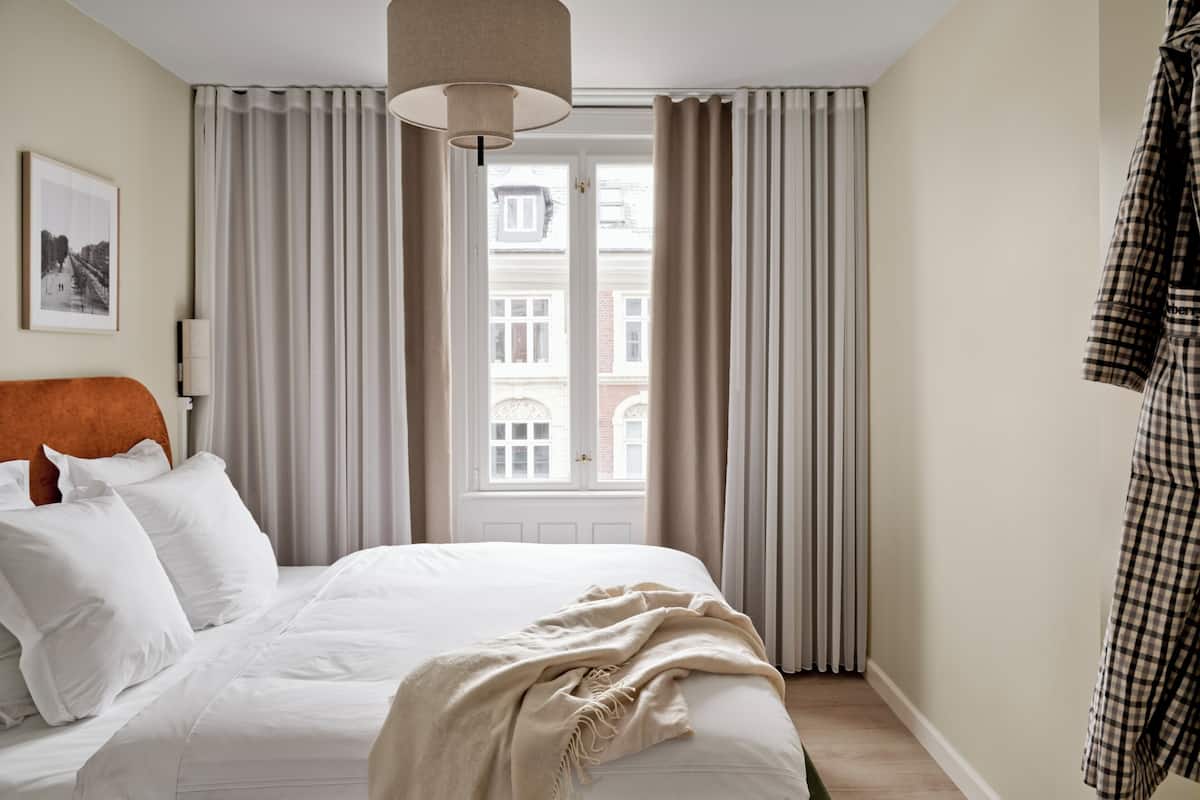 Een net opgemaakt bed met witte lakens en een beige deken, een groot raam met gordijnen, en een geruit kledingstuk aan de wand.
