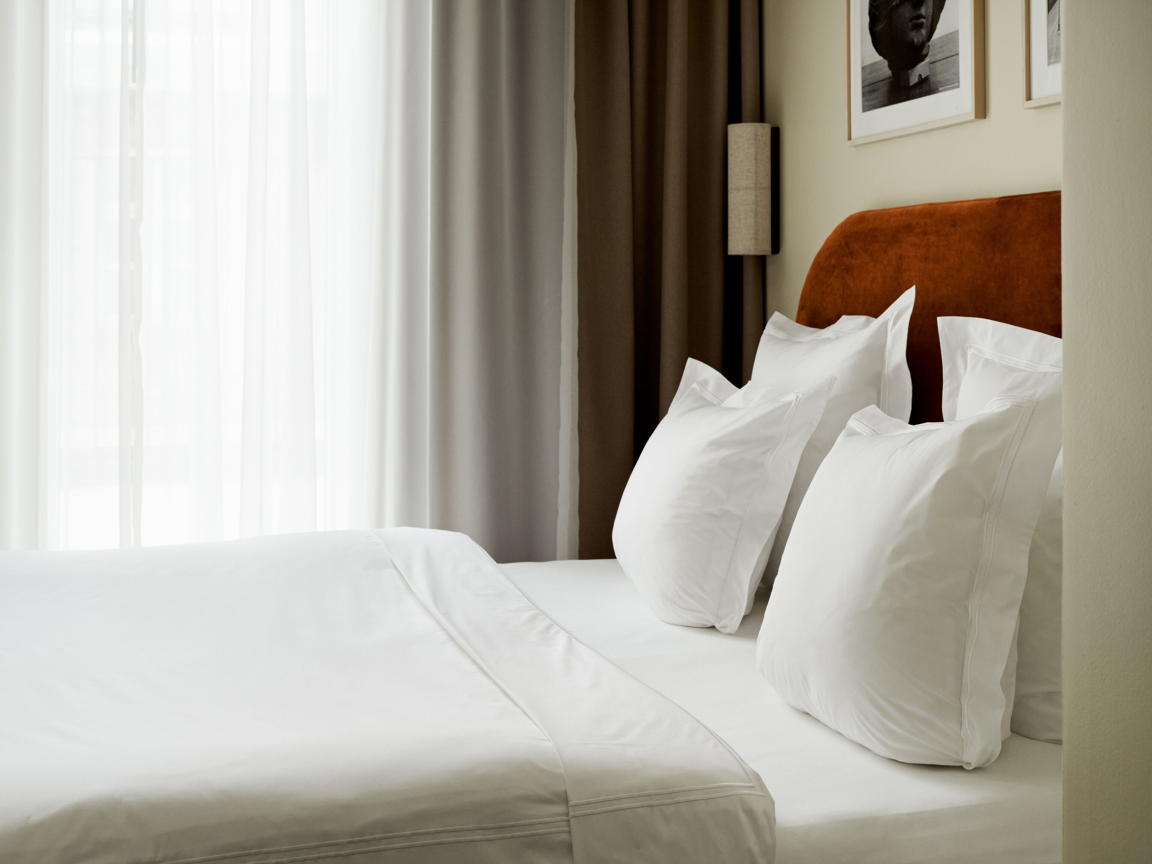 Una cama bien tendida con sábanas y almohadas blancas, cabecera de madera y un ventanal con cortinas traslúcidas.