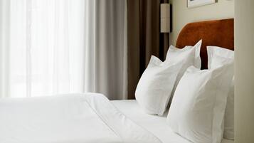 Una cama bien tendida con sábanas y almohadas blancas, cabecera de madera y un ventanal con cortinas traslúcidas.