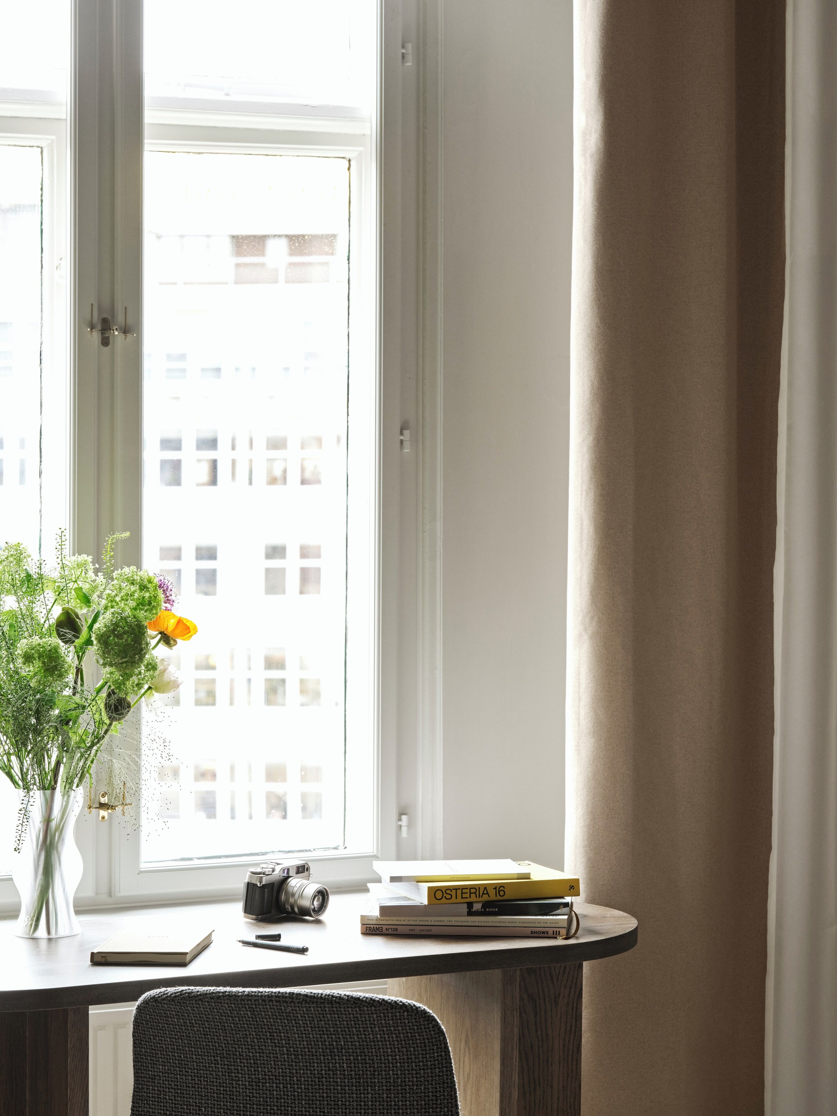 A room with a desk by a window, a vase with flowers, a camera, and books.