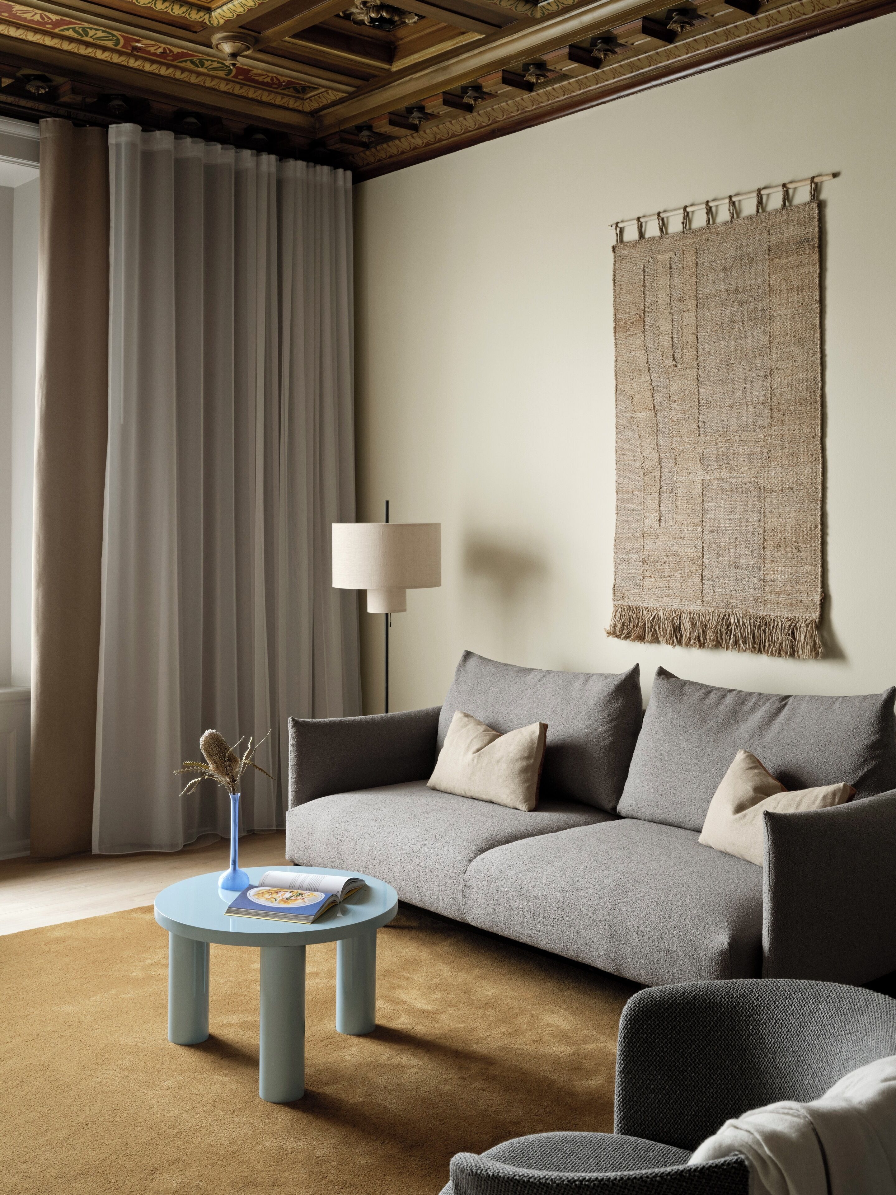 Un salon moderne avec un canapé gris, une table basse ronde et une grande tapisserie tissée accrochée au mur.