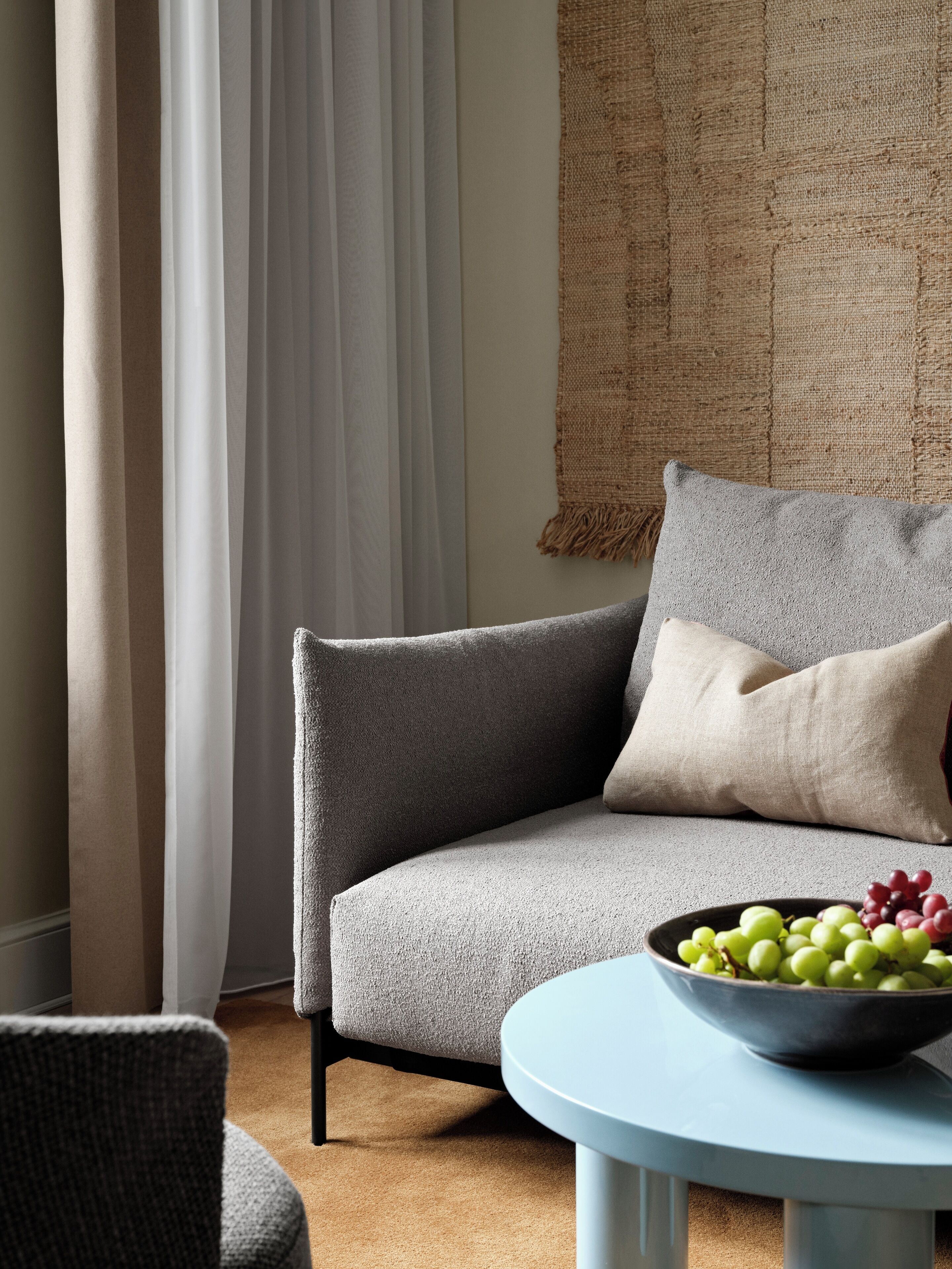 A modern living room with a grey sofa, a round table with a bowl of fruit, and a woven tapestry on the wall.