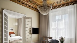 A spacious hotel room with a large bed, a television, a chandelier, and a view of the city through sheer curtains.
