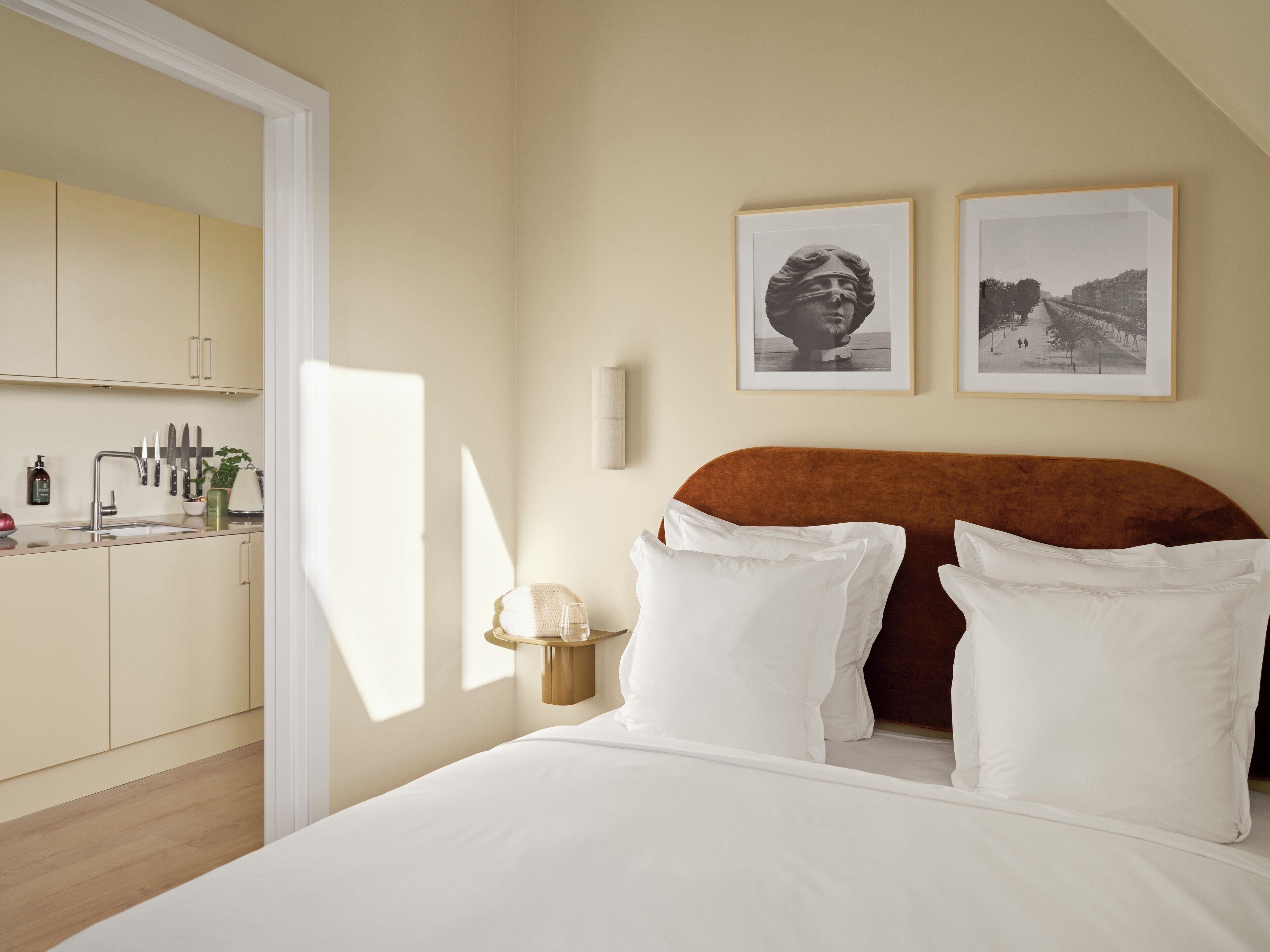 A bedroom with a bed, pillows, a nightstand, a lamp, a framed picture, and a view of a kitchen.